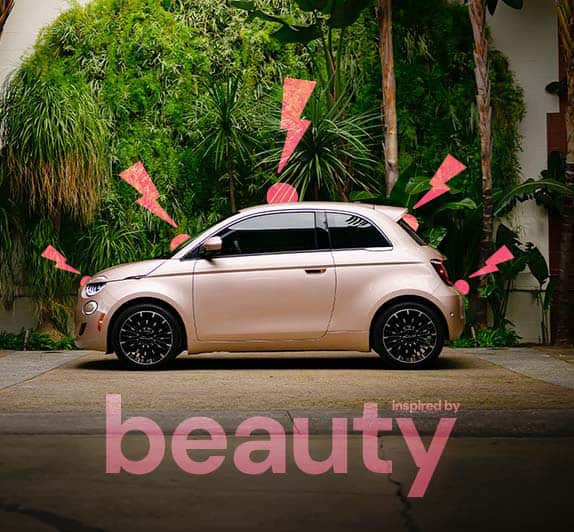 A side angle of the driver-side of a rose gold 2025 Fiat 500e Inspired by Beauty, parked on the street next to trees and plants, with illustrated hot pink zigzagged exclamation points in the right side foreground.
